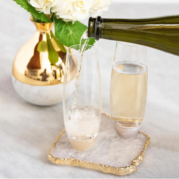 A bottle pours champagne into an Elevo Champagne Smoke Agate Glass by Anna New York, beside another filled glass. Both are on decorative coasters, next to a marble vase with white flowers and green leaves on a light surface.