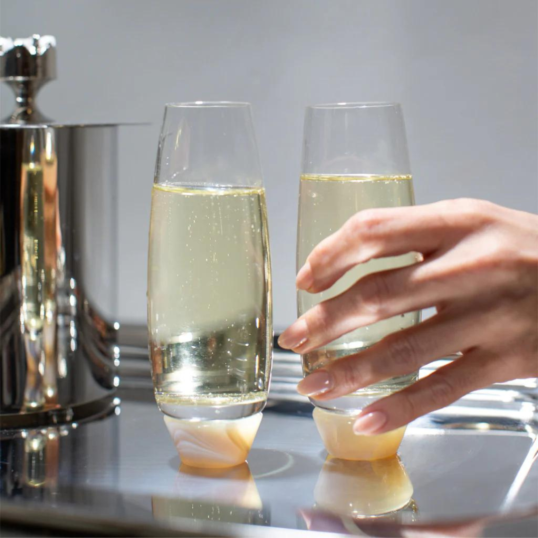 A hand reaches for an Elevo Champagne Smoke Agate Glass by Anna New York from a set of two, both crafted from lead-free crystal with unique angled bases, resting on a reflective surface.