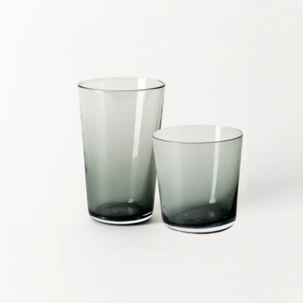 Two translucent charcoal hand-blown glass tumblers—one tall, one short—from Gary Bodker's Everyday Glasses collection are placed side by side on a plain white background.