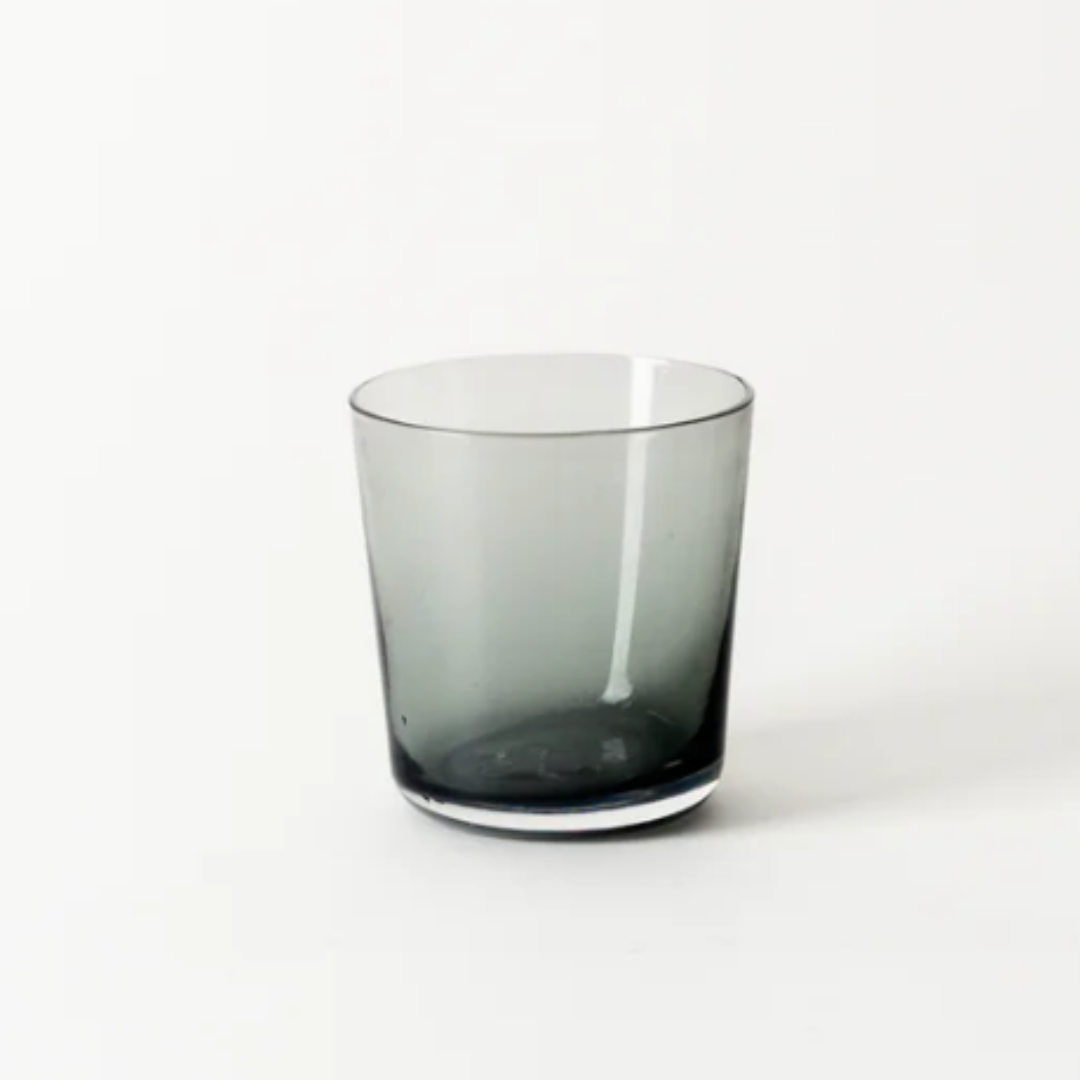 The Everyday Glasses Charcoal by Gary Bodker is a smoky gray, transparent, hand-blown glass tumbler with a simple cylindrical shape, elegantly displayed against a plain white background.