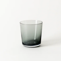 The Everyday Glasses Charcoal by Gary Bodker is a smoky gray, transparent, hand-blown glass tumbler with a simple cylindrical shape, elegantly displayed against a plain white background.