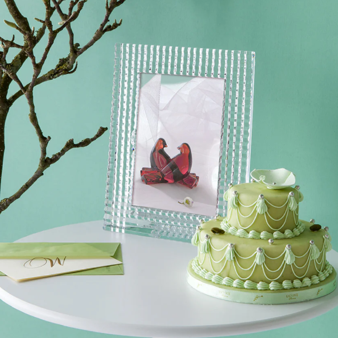 A round white table holds a green tiered cake with white icing, a yellow-green envelope, and Baccarat's Eye Photo Frame displaying two red glass birds. A leafless branch extends from the left against a mint-green wall.