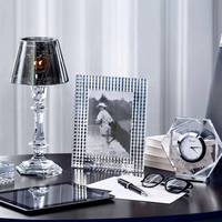A stylish desk setup featuring the Baccarat Eye Photo Frame, a crystal lamp, faceted crystal clock, stacked books, glasses, a tablet, and stationery arranged neatly on a dark surface.