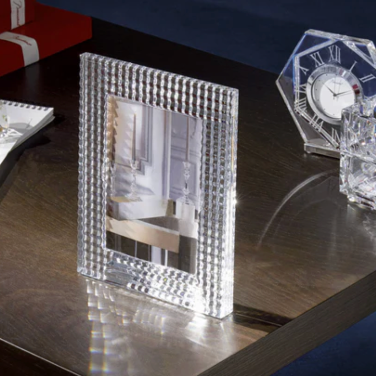 A Baccarat Eye Photo Frame and a glass clock by Nicolas Triboulot rest on a dark wooden table, reflecting light. In the background, a red box and notepad are partly visible.
