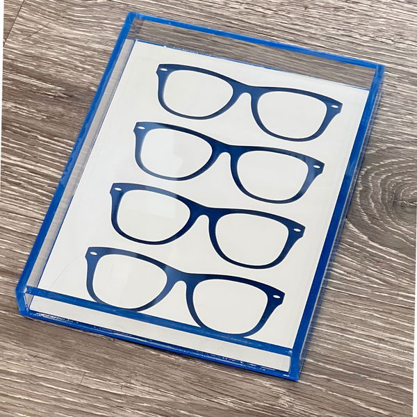 The Eye Candy Specs Eyeglass Tray is a rectangular luxury tray with a blue border and three pairs of illustrated blue eyeglasses on a white background, shown on a wood-textured surface.