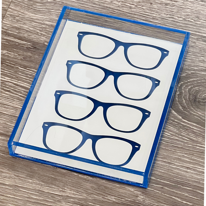 The Eye Candy Specs Eyeglass Tray is a rectangular luxury tray with a blue border and three pairs of illustrated blue eyeglasses on a white background, shown on a wood-textured surface.