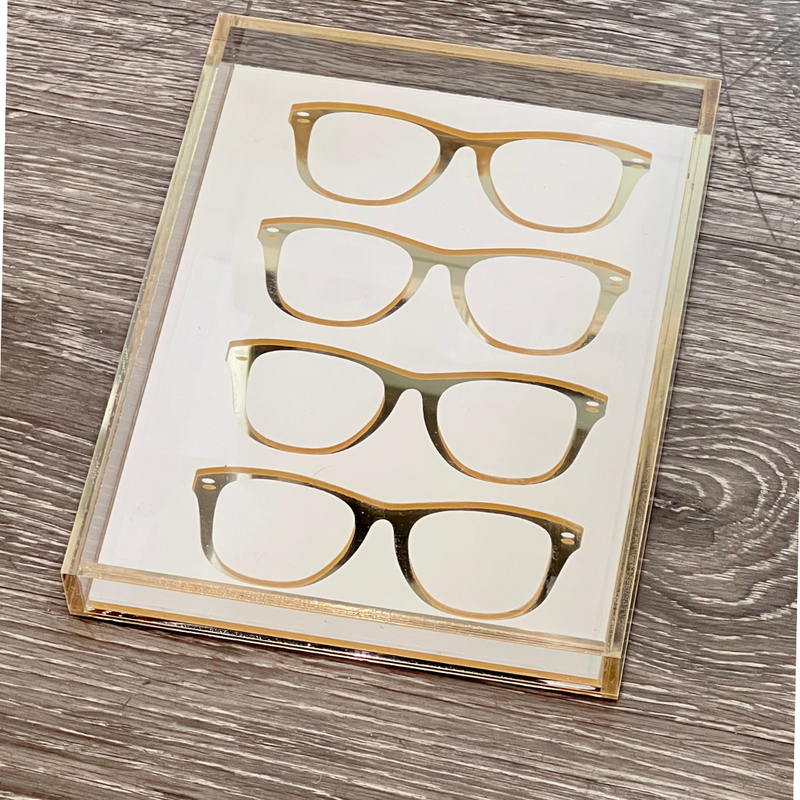 The Eye Candy Specs Eyeglass Tray features four gold eyeglass frame silhouettes, vertically and evenly spaced, displayed within a clear acrylic holder on a wooden surface.