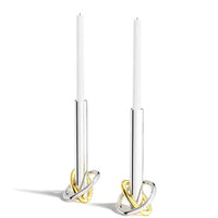 The Anna New York Ring Candle Holder Set of 2 features stainless steel holders with intertwined gold and silver bases, topped with white taper candles—an elegant accent for any modern decor.