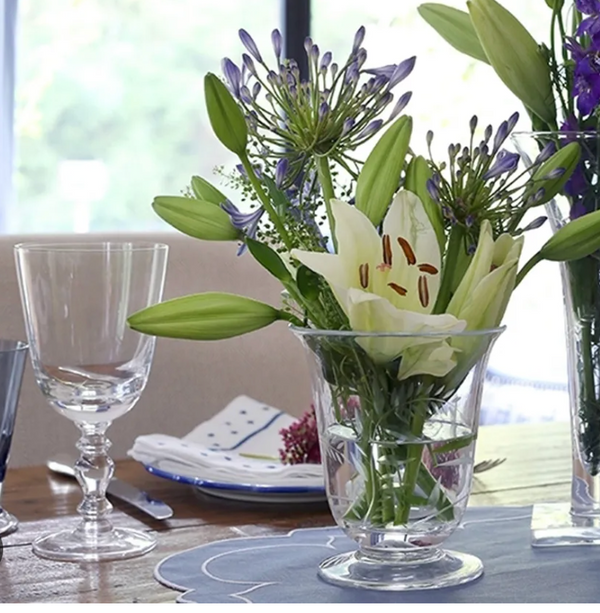 A set dining table features William Yeoward's Fanny Country Drinkware, a handmade crystal wine goblet beside a clear glass vase with white lilies and purple flowers, glassware, plates, and a blue placemat. The background is softly blurred.