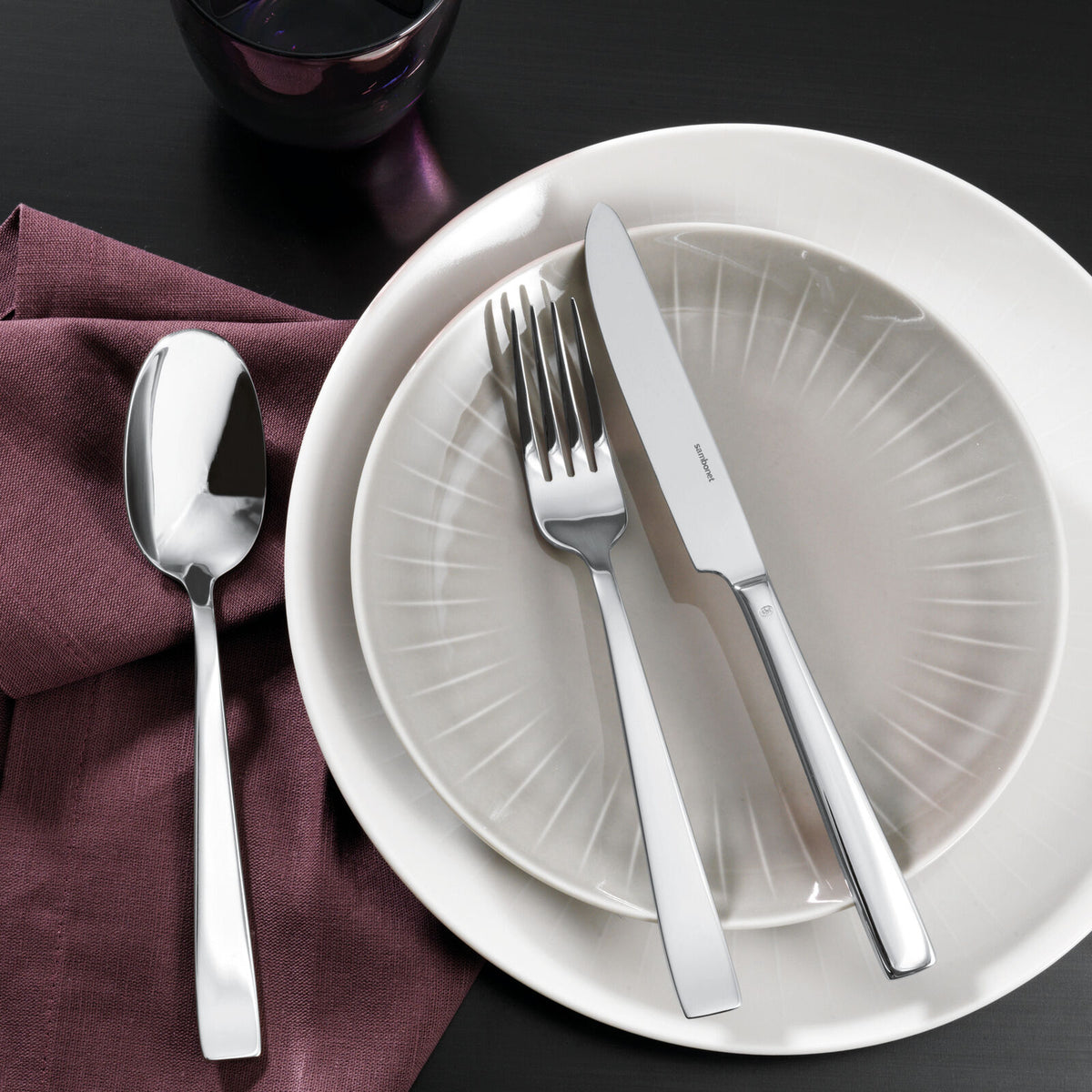 A neatly arranged table features a white and gray plate, Sambonet Flat Flatware 5 Piece Setting in stainless steel with a mirror finish, a spoon on a purple napkin to the left, and a dark glass above—all set on a black surface.