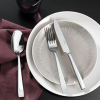 A neatly arranged table features a white and gray plate, Sambonet Flat Flatware 5 Piece Setting in stainless steel with a mirror finish, a spoon on a purple napkin to the left, and a dark glass above—all set on a black surface.