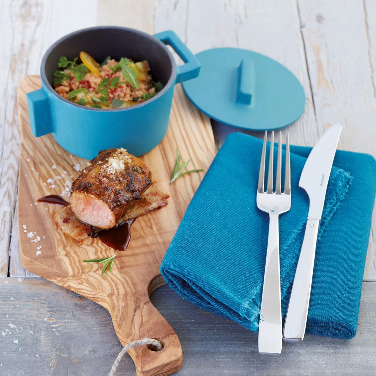 A serving board with sauced meat, a blue pot of grain salad, and the Sambonet Flat Flatware 5 Piece Setting in stainless steel with a mirror finish, elegantly arranged on a folded blue napkin beside the board.