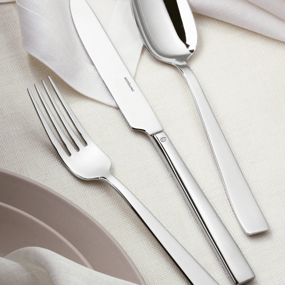 The Sambonet Flat Flatware 5 Piece Setting in stainless steel, featuring a mirror-effect finish, is arranged on a cream tablecloth beside folded white napkins and light beige plates.