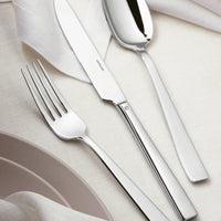 The Sambonet Flat Flatware 5 Piece Setting in stainless steel, featuring a mirror-effect finish, is arranged on a cream tablecloth beside folded white napkins and light beige plates.