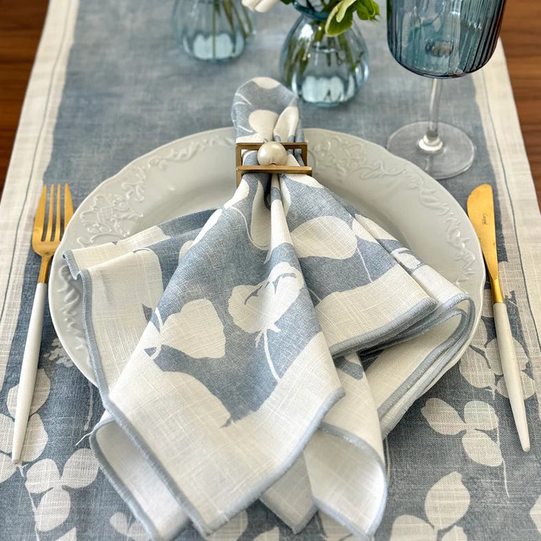 A table setting with a white plate on blue and white floral linens, gold and white cutlery, and a Bodrum Fleur de Lin Napkin (Set of 4 - Blue) folded with a ring. A blue glass and a vase of white flowers are in the background.
