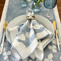A table setting with a white plate on blue and white floral linens, gold and white cutlery, and a Bodrum Fleur de Lin Napkin (Set of 4 - Blue) folded with a ring. A blue glass and a vase of white flowers are in the background.