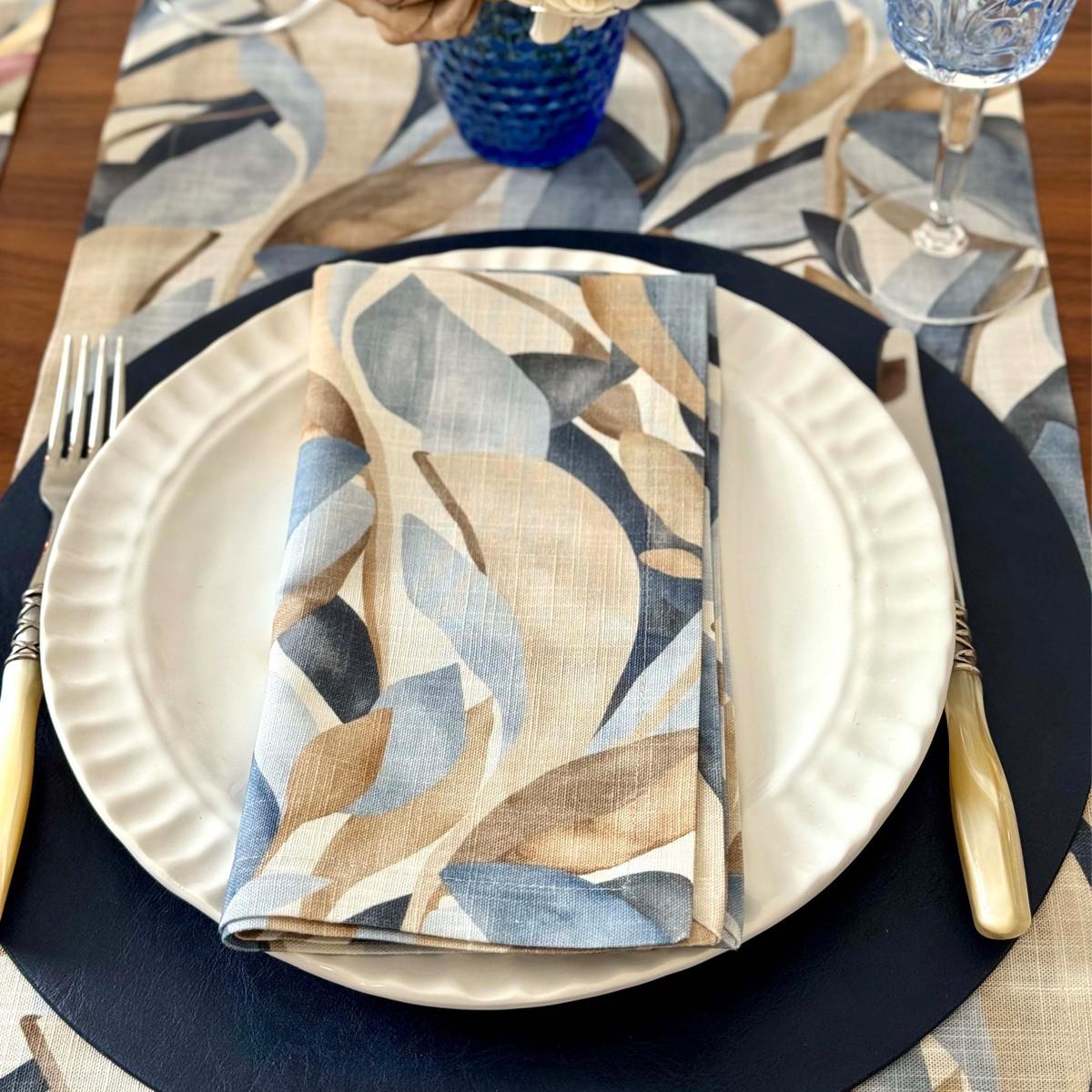 A table setting featuring Bodrum's Flora Napkin Set of 4, with a blue, beige, and brown leaf pattern that complements the fall tablescape, paired with a white plate on a dark placemat, light-handled cutlery, and a matching table runner.