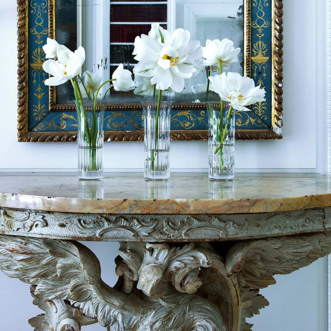 Three clear Flora Vases by Baccarat with white flowers adorn an ornate marble-topped table, showcasing exquisite craftsmanship in home décor, as a decorative framed mirror on the wall reflects their timeless elegance.