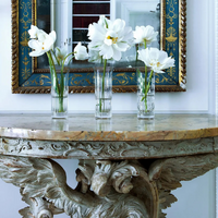 Three clear Flora Vases by Baccarat with white flowers adorn an ornate marble-topped table, showcasing exquisite craftsmanship in home décor, as a decorative framed mirror on the wall reflects their timeless elegance.