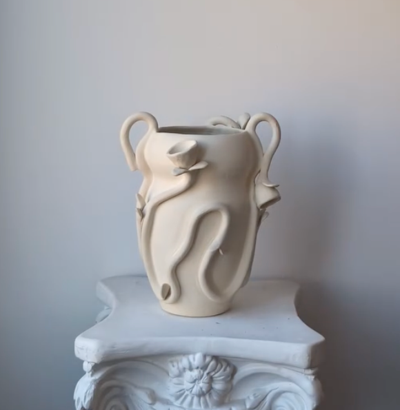 The Terroso Studios Floral Vase, a cream ceramic piece with abstract handles and wavy sculptural accents, sits on a white ornate pedestal against a simple light backdrop.