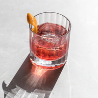 A red cocktail with a large ice cube and orange peel garnish sits on a light surface in an elegant Fluted Double Old Fashioned glass by Richard Brendon, casting a shadow.