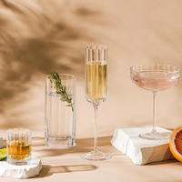 Cocktails are served in elegant glassware on a beige surface, including whiskey, a highball with rosemary, and the Richard Brendon Fluted Champagne Glass. Grapefruit slices and lime add vibrant garnishes to the display.