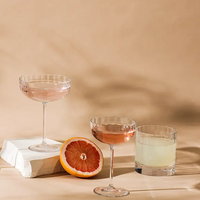 Three Richard Brendon Fluted Coupe glasses, crafted from mouth-blown glass, hold pink and clear drinks on a beige surface with a halved grapefruit, creating an elegant display against a neutral backdrop.