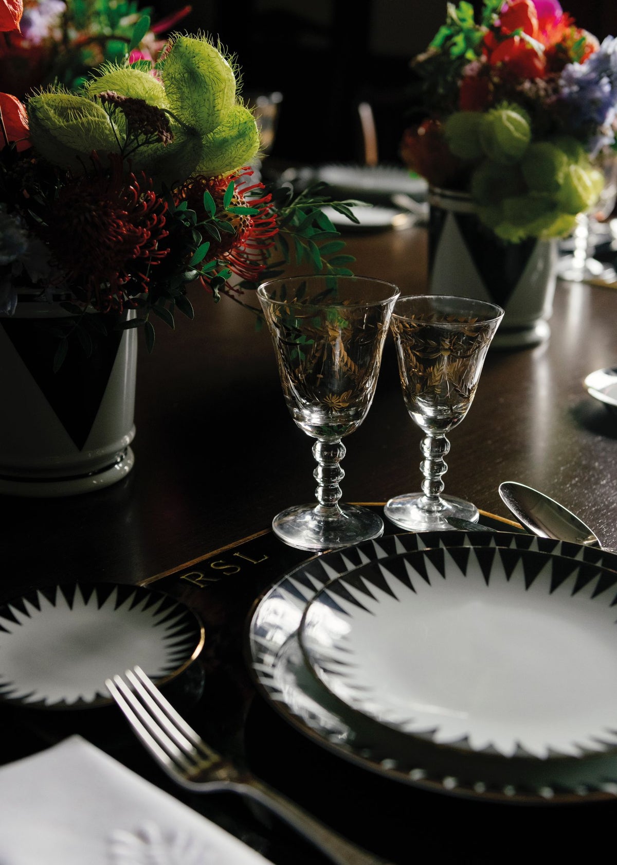 A close-up of an elegant table setting featuring Molecot’s Punk Dinnerware, two ornate wine glasses, silver cutlery, and floral centerpieces in white vases on a dark wooden table.