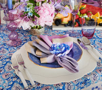 A vibrant table features a lavender and cream napkin with a blue flower napkin ring on a blue plate, Kim Seybert Mirage Flatware 5 pc Place Set in White/Silver (dishwasher safe), glassware, purple goblets, floral centerpiece, and fresh fruit platter.