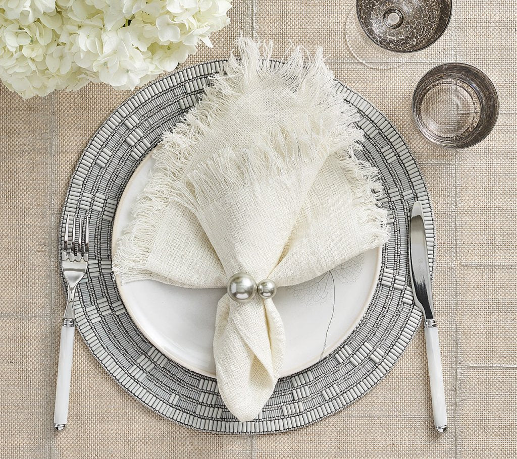 Create a farmhouse chic table with Kim Seybert's Fringe Napkin White & Silver Set of 4, paired with a silver-rimmed plate, white cutlery, two glasses, and a white floral centerpiece on a textured beige tablecloth.