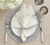 Create a farmhouse chic table with Kim Seybert's Fringe Napkin White & Silver Set of 4, paired with a silver-rimmed plate, white cutlery, two glasses, and a white floral centerpiece on a textured beige tablecloth.