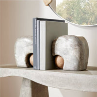 Two gray Melt Bookends Set of 2 by Arteriors, with marble bases and wooden spheres, support three books on a curved beige console table. Above, a round mirror reflects the trees outside.