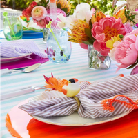 A vibrant outdoor table features striped napkins with bird napkin rings, floral centerpieces, bright plates, and Kim Seybert’s Gala Stemless Tumbler Set 4 adding sparkle to a blue-and-white striped tablecloth.