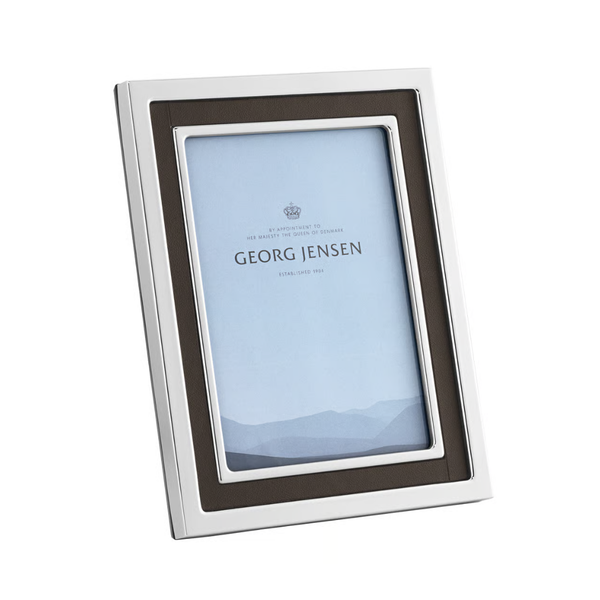 The Georg Jensen Manhattan Picture Frame features a silver and black stainless steel design, displaying a placeholder image with subtle mountains and the brand name. The frame stands elegantly at an angle.