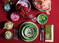 A table set on a red cloth features Ginori 1735 Oriente Italiano Dinnerware Malachite plates, bowls, and cups with intricate designs in green, plus silver cutlery, wine glasses, and a green napkin with a white border.