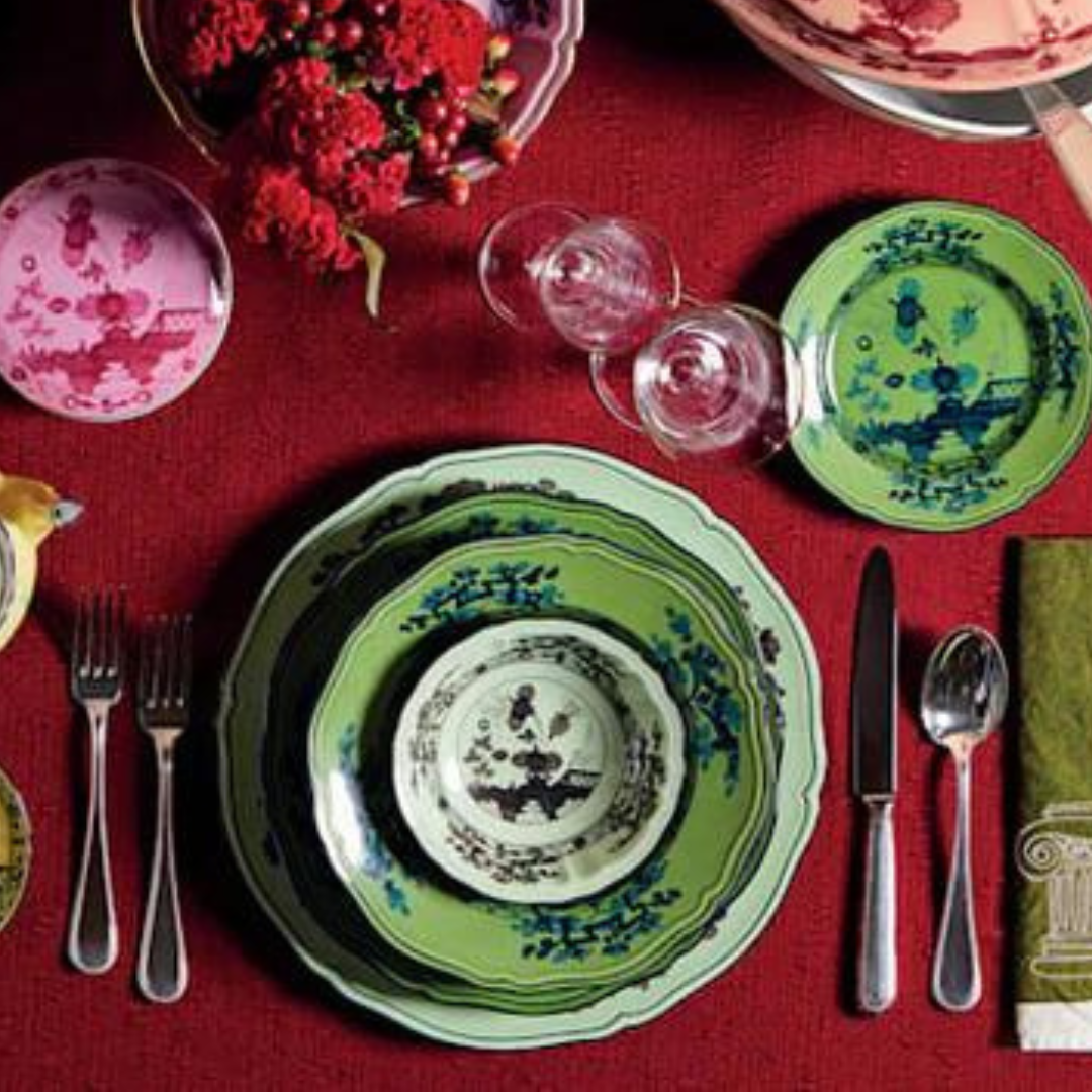 A formal table setting on a red tablecloth, featuring stacked Ginori 1735 Oriente Italiano Dinnerware Malachite plates, silverware, two clear wine glasses, a green napkin, and small pink and green floral decorative plates.