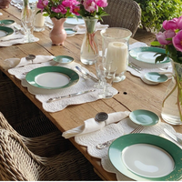 A rustic wooden table is set with Molecot's Golden Green Dinnerware, white napkins, silver cutlery, and glassware. Pink flowers in vases and white candles serve as centerpieces, while wicker chairs surround the table.