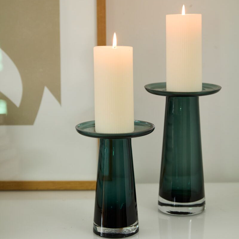 Two lit white pillar candles stand in dark green Accent Decor Topple Vase Green candle holders on a white surface, perfect for coffee table decor.
