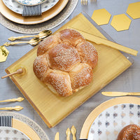 A round challah with sesame seeds rests on the Apeloig Solid Acrylic Challah Board & Knife. Gold cutlery, bee-themed plates, and a gray tablecloth with gold hexagons complete the modern table setting.