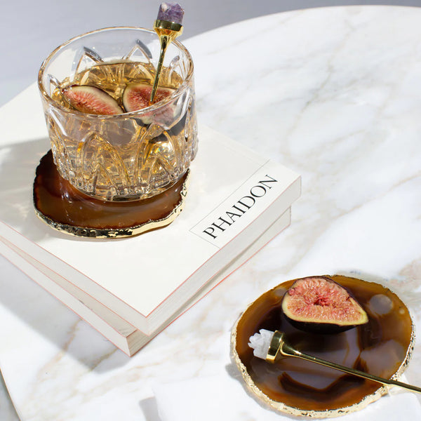 A crystal glass with a drink and fig slices rests on a brown agate coaster atop a white PHAIDON book. Nearby, an Anna New York Hospitality Cocktail Pick in gold & amethyst druze and a fig slice are arranged on a plate on the marble surface.