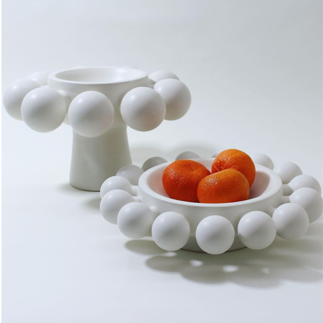 Two modern, white porcelain bowls from KleinReid’s Nimbus Halo Bowl collection feature round-rimmed accents; one holds oranges and the other, empty and elevated on a pedestal, are set against a plain white background.