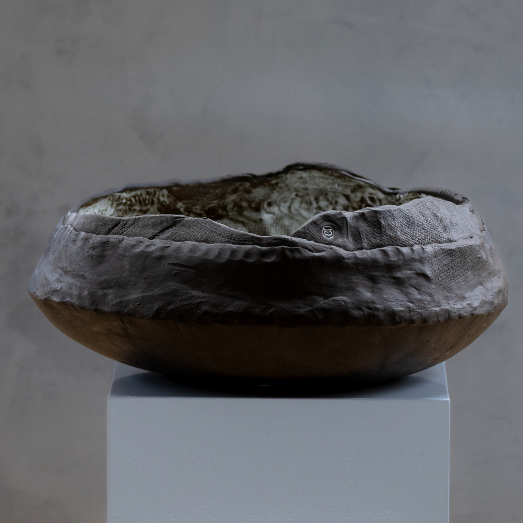 The Obsidian Wave Bowl by Kirti Sheoran Studio is a large, shallow ceramic piece made from black stoneware clay with an uneven, textured surface and earthy tones, displayed on a white pedestal against a gray background.