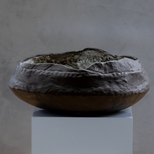 The Obsidian Wave Bowl by Kirti Sheoran Studio is a large, shallow ceramic piece made from black stoneware clay with an uneven, textured surface and earthy tones, displayed on a white pedestal against a gray background.