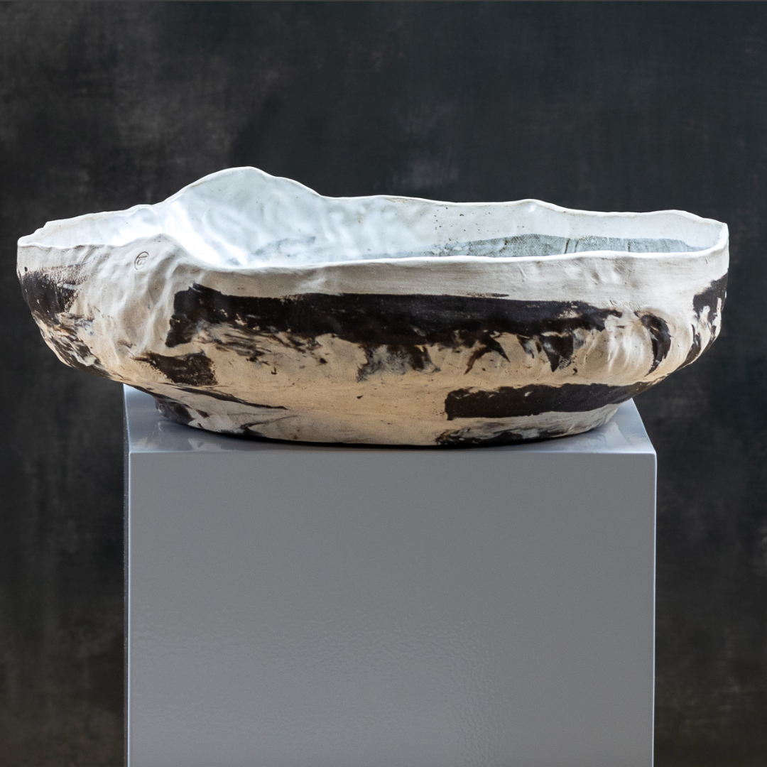 The Desert Basin Bowl by Kirti Sheoran Studio—a handmade, large ceramic bowl with an irregular wavy rim and bold dark brown patches on white glaze—adds artistic flair to modern decor, displayed on a white pedestal against a dark backdrop.