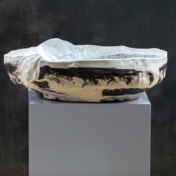 The Desert Basin Bowl by Kirti Sheoran Studio—a handmade, large ceramic bowl with an irregular wavy rim and bold dark brown patches on white glaze—adds artistic flair to modern decor, displayed on a white pedestal against a dark backdrop.