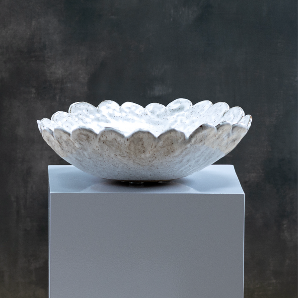 The Daisy Bowl by Kirti Sheoran Studio is a white, scalloped ceramic piece with a brown speckled finish, displayed on a gray pedestal—ideal as handmade functional ware for modern home decor.