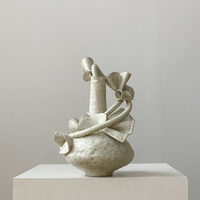 The Latch Ribbon White Vessel by Latch Key Ceramics features abstract organic forms and curved protrusions in speckled off-white ceramic, shown on a white pedestal with a light gray background.