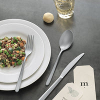 A plate of mixed vegetables and grains rests on stacked white dishes with a fork on top. Next to it are Sambonet's Hannah Antico Flatware 5 Piece Setting, a drink, a small bread roll, and paper cards on a gray tabletop.