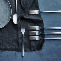 The Sambonet Hannah Antico Flatware 5 Piece Setting, featuring textured gray finishes, includes a modern stainless-steel spoon and four sleek forks arranged on a dark cloth over blue, with the forks neatly lined up on the right side.