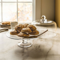 The Simon Pearce Hartland Cakeplate, expertly crafted by glassblowers, displays golden-brown pastries with a knife nearby on a marble counter. Sunlight streams through the window as its glass dome rests to the side.
